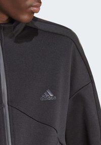 Melna sporta jaka ar gludu tekstūru, kas aprīkota ar rāvējslēdzēju un tonālā Adidas logo uz krūtīm. Redzami stūra dizaina elementi.