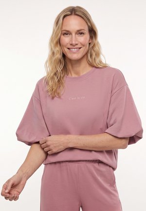 Glimlachende vrouw met golvend blond haar, draag een mauve T-shirt met korte mouwen en bijpassende broek, houdt met de ene hand haar andere arm vast.