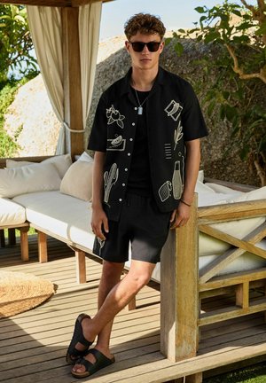 Young man wearing black printed short-sleeve shirt and shorts with sunglasses, leaning on wooden post on shaded deck with cushioned seating.