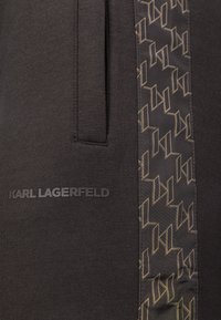 Ciemny materiał z pionowym wzorowanym paskiem, na którym znajduje się geometryczny motyw oraz dyskretne logo Karl Lagerfeld w pobliżu pionowej kieszeni.