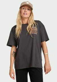 Blonde vrouw met een beige pet en een oversized zwart T-shirt met een oranje surfthema logo op de borst, staand tegen een effen achtergrond.