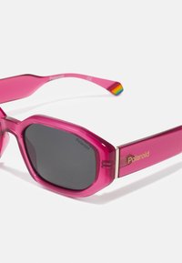 Gafas de sol translúcidas rosas con montura rectangular, lentes oscuros y un detalle dorado con la marca "Polaroid". Presenta un detalle arcoíris en las patillas.