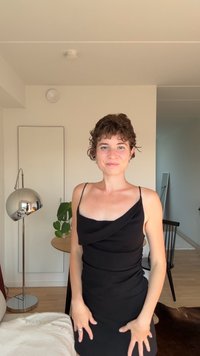 Sort kjole med draperet halsudskæring og tætsiddende silhuet. Glat stof, knælang. Baggrunden inkluderer et spejl, en lampe og indendørs planter.