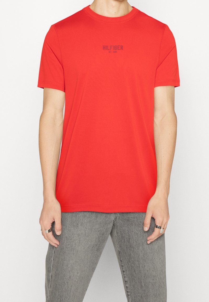 Homme portant un t-shirt rouge uni à manches courtes avec le texte « HILFIGER » sur la poitrine, associé à un jean en denim gris.