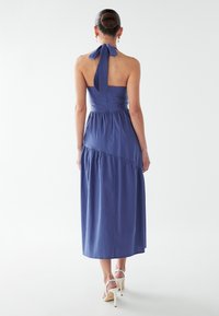 Modra obleka s trakovi okoli vratu, oprijetim zgornjim delom in večplastno krilom, okrašena s povezanim detajlom na hrbtu ter gladko, tekočo teksturo tkanine.