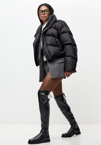 Mujer con chaqueta de plumas negra, pantalones cortos grises, botas de cuero negras hasta el muslo y gafas caminando frente a un fondo blanco liso.