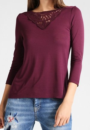 Femme portant un haut à manches longues bordeaux avec un détail en dentelle au niveau de l'encolure et un jean bleu avec une broderie florale sur la cuisse.