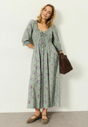 Frau in einem grünen, geblümten Midikleid mit gerafftem Oberteil und Puffärmeln, die eine braune Wildledertasche hält und braune Mary Jane Schuhe trägt.