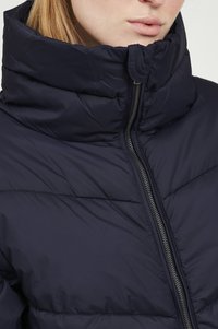 Veste matelassée bleu marine avec un col haut, fermeture éclair et texture matelassée, offrant une isolation avec un design moderne et épuré.