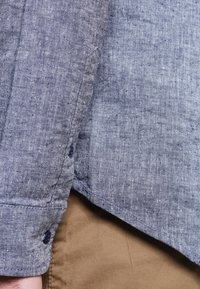 Camisa de lino en un tono azul oscuro con un acabado texturizado. Presenta un dobladillo redondeado y botones de material oscuro. Tela marrón visible por debajo.