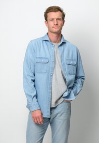Camisa de mezclilla azul claro con dos bolsillos en el pecho, cierre de botones y mangas largas sobre una camiseta gris, combinada con jeans claros.