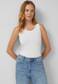 Mujer con cabello castaño claro vestida con una blusa blanca sin mangas y jeans azules, de pie con las manos detrás de la espalda contra una pared gris.