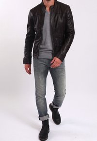 Brun läderjacka med en figursydd design, dragkedjestängning och subtila axeldetaljer, bärs över en grå V-ringad tröja och blå jeans.