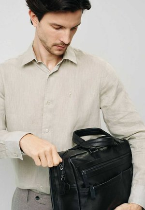 Czarna skórzana torba na laptopa z wieloma zamykanymi na zamek przegródkami, elegancki design oraz mocne rączki u góry. Na sobie noszona jest jasnobeżowa lniana koszula.