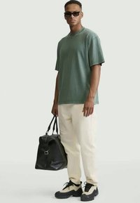 T-shirt verde di cotone, modello oversize, abbinato a pantaloni crema e scarpe da ginnastica nere. Tiene una borsa a mano in pelle nera.