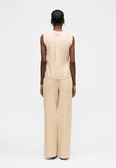 Femme aux cheveux courts portant un haut beige sans manches et un pantalon à jambes larges, debout de dos contre un fond uni.