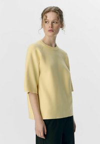 Haut jaune clair à manches courtes avec un col rond, texture lisse et coupe décontractée, associé à un pantalon noir sur fond neutre.