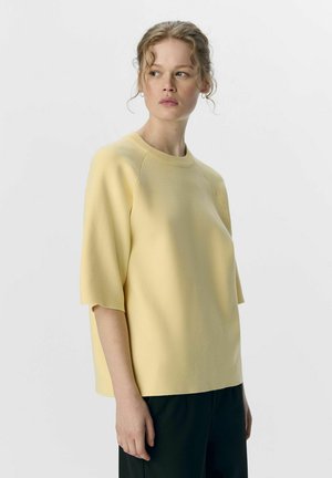 Haut jaune clair à manches courtes avec un col rond, texture lisse et coupe décontractée, associé à un pantalon noir sur fond neutre.