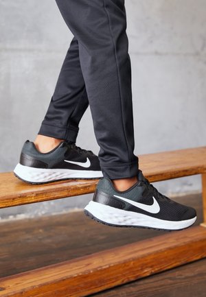 Baskets de sport noires avec le swoosh blanc de Nike, tige en mesh, talon texturé et épaisse semelle extérieure blanche, portées avec un jogging noir ajusté sur une marche en bois.