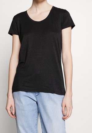 Osoba ubrana w czarną koszulkę z krótkim rękawem i jasnoniebieskie dżinsy, stojąca na tle jednolitego jasnego tła.