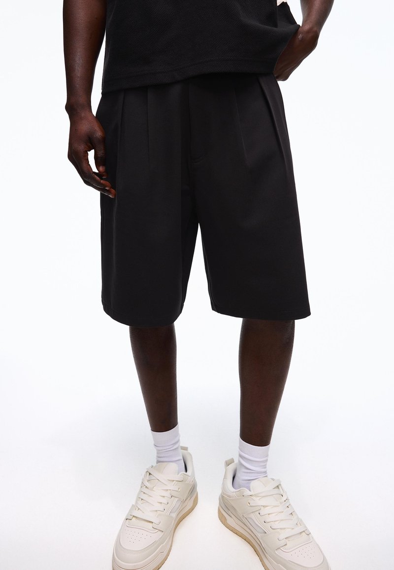 Homme portant un short ample noir, des chaussettes blanches mi-mollet et des baskets écru, sur un fond uni.