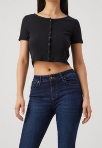 Schwarzes kurzärmeliges, verkürztes Oberteil mit Knöpfen an der Vorderseite, kombiniert mit dunkelblauen Jeans in enger Passform und frontalen Taschen.