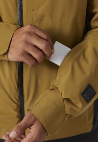 Veste imperméable couleur kaki avec une fermeture éclair noire ; comporte un patch avec le logo de la marque. Main insérant une carte dans une poche à fermeture éclair.