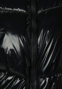 Černá vatovaná bunda s lesklým povrchem, vertikálním zapínáním na zip a prošíváním. Textura je hladká s polstrovanými částmi.
