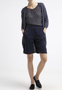 Mörkblå shorts med elastisk midja i kombination med en prickig topp och en lång, öppenstickad cardigan i en mörk nyans. Enkla svarta foppatofflor.