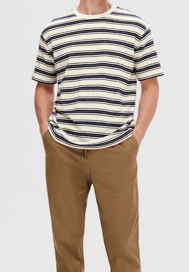 Beżowa koszulka z krótkim rękawem w paski, z granatowymi paskami, okrągłym dekoltem i luźnym krojem, połączona z beżowymi spodniami. Wykonana z miękkiego materiału bawełnianego.