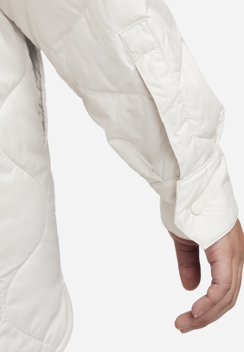 Manche de veste matelassée blanche avec une texture lisse, dotée d'une fermeture à bouton-pression et d'une poche visible sur le bras.