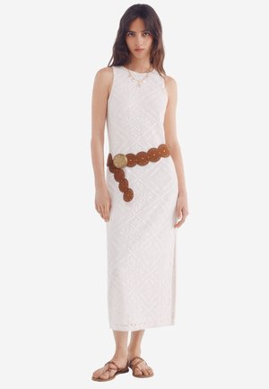 Weißes Spitzen-Maxikleid mit geometrischem Muster, ausgestattet mit einem braunen Gürtel mit runden Akzenten. Sandalen mit überkreuzten Riemen.