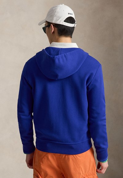 Homme portant un sweat à capuche bleu roi, un pantalon orange, une casquette Polo blanche et des lunettes de soleil foncées, de dos sur un fond uni.
