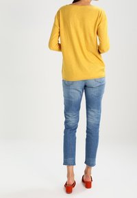 Gelbes langärmeliges Strickoberteil mit lockerer Passform, kombiniert mit hellblauen Slim-Fit-Jeans und roten Absatzsandalen.