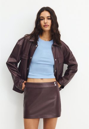 Veste en faux cuir marron avec des boutons-pression, assortie à un haut ajusté bleu clair et une jupe mini en faux cuir marron coordonnée.