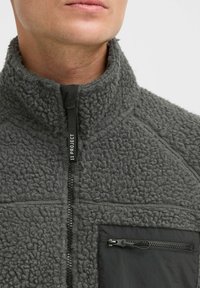 Grauer Fleece-Halbzip-Pullover mit hohem Kragen, strukturiertem Oberflächenmaterial und einer schwarzen, mit Reißverschluss versehenen Brusttasche; zeichnet sich durch kontrastierende Materialien aus.