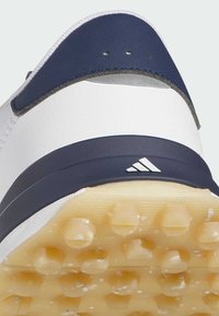 Gros plan sur le talon d’une chaussure de sport blanche et bleu marine avec des crampons jaunes texturés sur la semelle et un petit logo Adidas sur la partie talon.