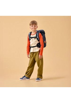 Young boy standing, wearing khaki pants, orange and cream long-sleeve shirt, blue sneakers, and a dark blue backpack with chest straps.