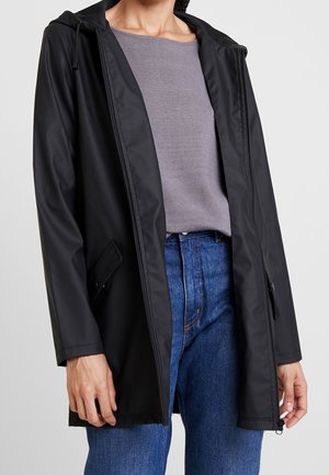 Person iført en sort hætte-regnjakke over en grå sweater og blå jeans, hvor jakken er oplyst og hænderne afslappede ved siderne.