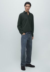 Dark green long-sleeve polo shirt with a collar, paired with grey straight-leg jeans and black shoes. Simple, textured fabric.