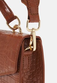 Coin de sac à main en cuir marron texturé avec des fermoirs métalliques dorés attachant la bandoulière au sac sur fond blanc.