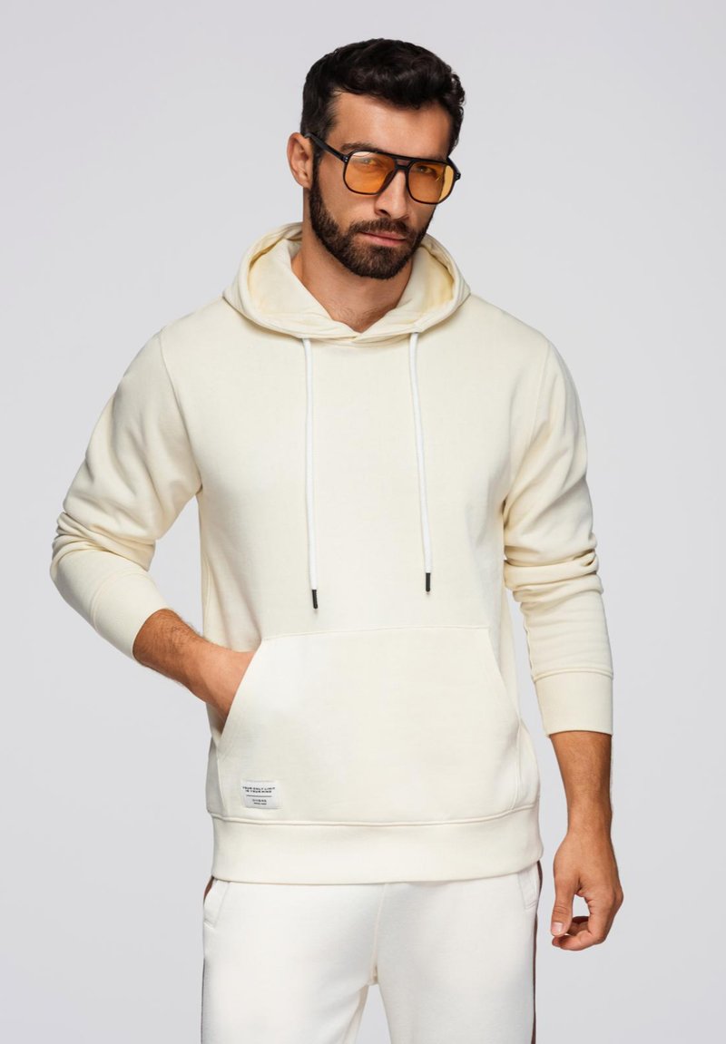 Crème-kleurige hoodie gemaakt van zachte stof, met een voorzak, witte trekkoorden en geribbelde boorden. Lijkt een relaxte pasvorm te hebben.