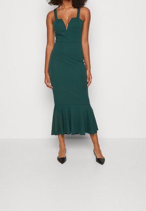 Kobieta w ciemnozielonej sukience midi bez rękawów z falbanką na dole, w czarnych butach z noskiem w szpic, stojąca na tle jednolitego białego tła.