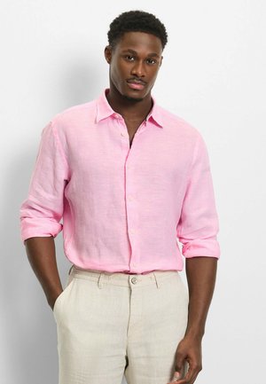 Junger Mann mit kurzen lockigen Haaren, der ein hellrosa Hemd mit hochgekrempelten Ärmeln und beige Hose trägt, vor einem schlichten Hintergrund steht.