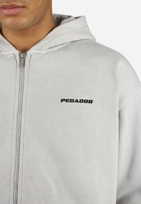 Sudadera gris con cremallera, hecha de un tejido suave, con capucha ajustable y el logo negro "PEGADOR" en el pecho. Diseño y textura simples.