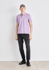 Svetlofialová polo košeľa s golierom, čierne slim-fit džínsy a čierne topánky. Model má tiež na sebe šedú čiapku. Jednoduché pozadie.