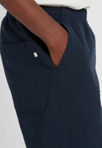 Navyblaue, strukturierte Jogging-Shorts mit elastischem Bund und Seitentasche, ausgestattet mit einem kleinen weißen Markentag an der Naht der Tasche.