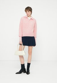 Giovane che indossa un maglione rosa, una gonna plissettata blu navy, scarpe nere con calzini scuri, mentre tiene una borsa bianca su uno sfondo bianco.