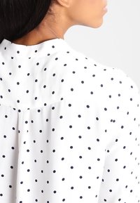 Vista dalla parte posteriore di una donna, con la parte superiore della schiena e il collo visibili. Indossa una blusa bianca con pois neri distribuiti uniformemente e ha i capelli raccolti, girata di spalle.