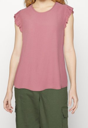 Kobieta ubrana w różowy top bez rękawów z falbankami na ramionach oraz oliwkowe spodnie typu cargo, stojąca na białym tle.